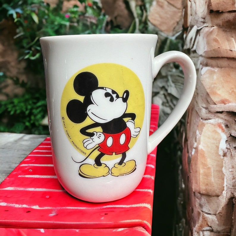 Peut inclure: Une tasse en c&eacute;ramique blanche avec un fond jaune et une image de dessin anim&eacute; de Mickey Mouse dans une tenue rouge et noire. La tasse a une poign&eacute;e noire et le texte "&copy; WALT DISNEY PRODUCTIONS" est imprim&eacute; sur le c&ocirc;t&eacute;.