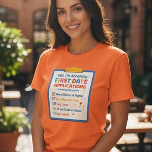 May include: Orange t-shirt with the text "Yes, I'm Accepting First Date Applications." The shirt lists requirements including a good sense of humor, the ability to plan a date, being a dog lover, and having great taste in music.