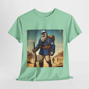 Puede incluir: Una camiseta verde con una ilustración vintage de una gran criatura peluda que lleva un uniforme militar francés y lleva un rifle. La criatura está de pie en un paisaje desértico con otros soldados en el fondo.