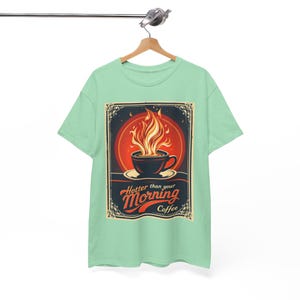 Op de afbeelding: Een lichtgroen T-shirt met een vintage-stijl grafische afbeelding van een koffiekopje met vlammen die eruit komen. De afbeelding zegt "Hotter than your Morning Coffee".