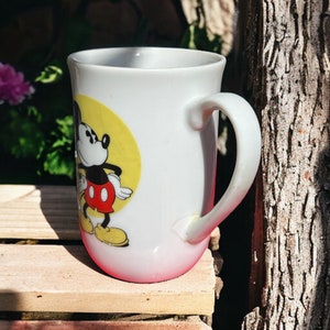 Peut inclure: Une tasse en c&eacute;ramique blanche avec un motif Mickey Mouse rouge et jaune. La tasse a une anse et un bord rose.