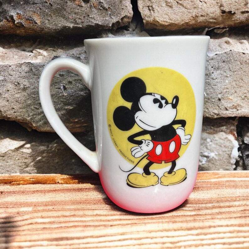 Peut inclure: Une tasse en c&eacute;ramique blanche avec un cercle jaune et une image de dessin anim&eacute; de Mickey Mouse en noir, rouge et jaune. Le texte "&copy; WALT DISNEY PRODUCTIONS" est imprim&eacute; sur la tasse.