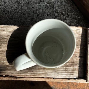Peut inclure: Une tasse en c&eacute;ramique blanche avec une anse. La tasse est vide et repose sur une surface en bois.