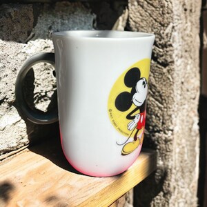 Peut inclure: Une tasse en c&eacute;ramique blanche avec une illustration de Mickey Mouse en jaune, rouge et noir. La tasse a une poign&eacute;e noire et le texte "&copy; WALT DISNEY PRODUCTIONS" est imprim&eacute; sur le c&ocirc;t&eacute;.