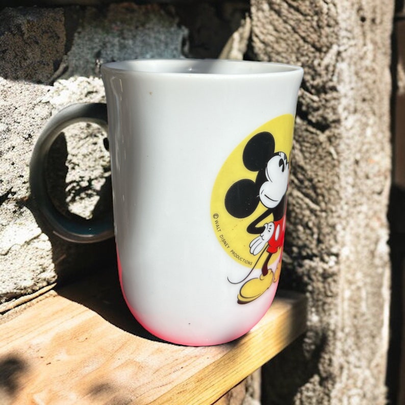 Peut inclure: Une tasse en c&eacute;ramique blanche avec une illustration de Mickey Mouse en jaune, rouge et noir. La tasse a une poign&eacute;e noire et le texte "&copy; WALT DISNEY PRODUCTIONS" est imprim&eacute; sur le c&ocirc;t&eacute;.