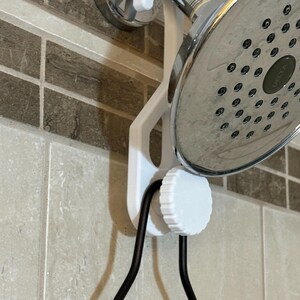 May include: A white shower head holder is mounted on a tiled wall, supporting a chrome shower head. The holder has a circular adjustment knob and a curved design to hold the shower head and hose. The black hose is visible.
