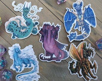 May include: A collection of fantasy dragon stickers in various colors, including teal, blue, purple, and orange. The stickers feature detailed illustrations of different dragon species, with some surrounded by clouds or water-like elements. Dice are scattered around the stickers.