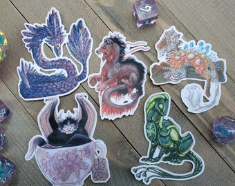 May include: A collection of fantasy creature stickers, including a blue dragon, a red and black beast, a spotted orange and brown creature, a green lizard-like figure, and a bat-like creature in a teacup. The stickers are on a wooden surface.