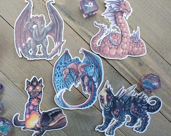 May include: A collection of fantasy-themed stickers featuring various dragon designs in shades of red, blue, and purple. The stickers are placed on a wooden surface, with several dice scattered around.