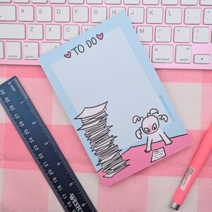 May include: A light blue to-do list notepad with a pink border and a cartoon illustration of a creature with a stack of papers. The notepad has the text "♡TO DO♡" at the top.