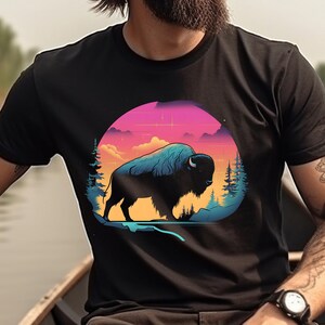May include: A black t-shirt with a graphic of a silhouette of a bison in front of a sunset with pink, orange, and blue colors. The bison is in a circle with trees on either side.