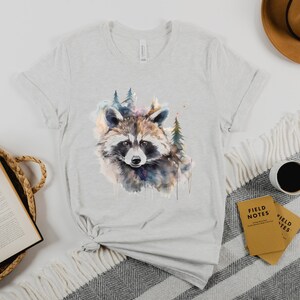 Puede incluir: Camiseta gris claro con una ilustraci&oacute;n de acuarela de un mapache y un bosque. El mapache tiene pelaje marr&oacute;n, gris y blanco. La camiseta est&aacute; hecha de material suave y es adecuada para ropa casual.