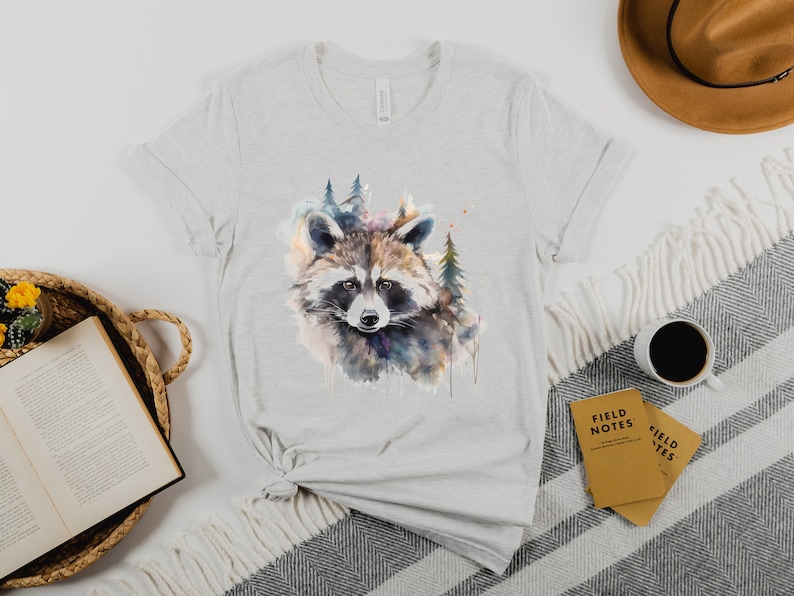 Puede incluir: Camiseta gris claro con una ilustraci&oacute;n de acuarela de un mapache y un bosque. El mapache tiene pelaje marr&oacute;n, gris y blanco. La camiseta est&aacute; hecha de material suave y es adecuada para ropa casual.