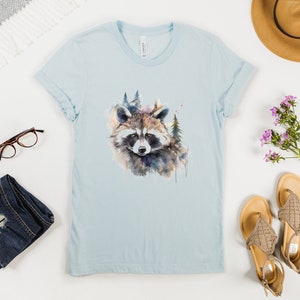 Puede incluir: Camiseta azul claro con un dise&ntilde;o de mapache en acuarela y elementos forestales. La camiseta est&aacute; colocada plana con las mangas dobladas, junto con accesorios como gafas, vaqueros, sandalias y un sombrero. El tema es la naturaleza y la ropa casual.