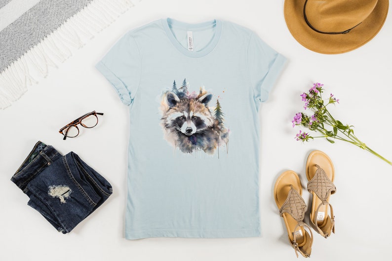 Puede incluir: Camiseta azul claro con un dise&ntilde;o de mapache en acuarela y elementos forestales. La camiseta est&aacute; colocada plana con las mangas dobladas, junto con accesorios como gafas, vaqueros, sandalias y un sombrero. El tema es la naturaleza y la ropa casual.
