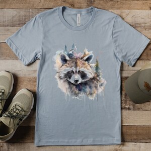 Puede incluir: Camiseta azul claro con un dise&ntilde;o de mapache en acuarela y elementos forestales. El mapache tiene una cara detallada con una m&aacute;scara negra y est&aacute; rodeado de pinceladas coloridas. Zapatillas de deporte beige y un sombrero verde tambi&eacute;n est&aacute;n en la imagen.