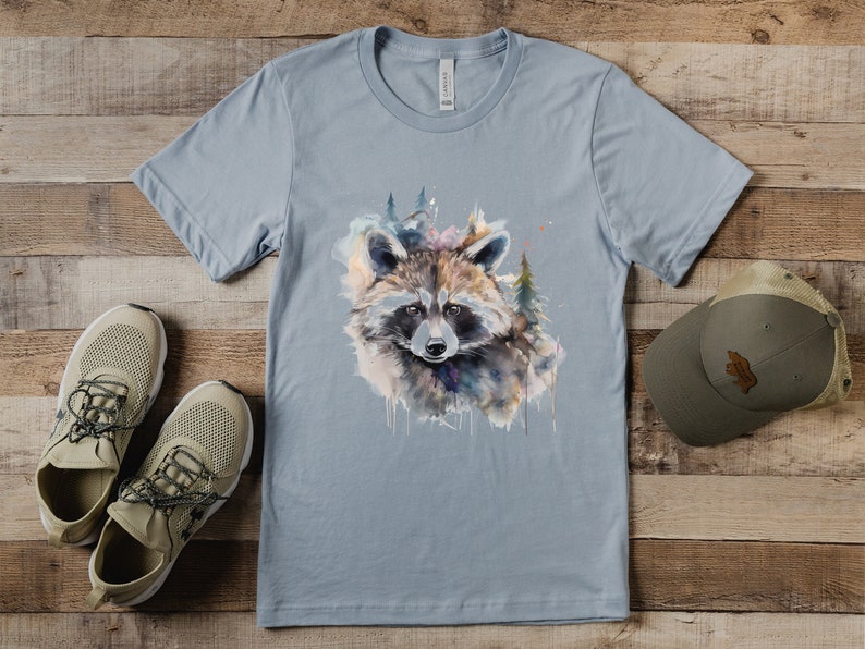 Puede incluir: Camiseta azul claro con un dise&ntilde;o de mapache en acuarela y elementos forestales. El mapache tiene una cara detallada con una m&aacute;scara negra y est&aacute; rodeado de pinceladas coloridas. Zapatillas de deporte beige y un sombrero verde tambi&eacute;n est&aacute;n en la imagen.