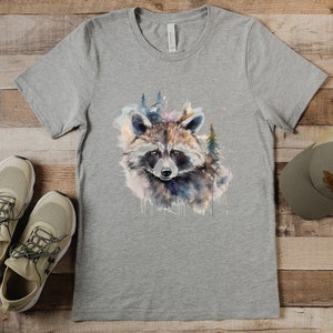 Puede incluir: Una camiseta gris con un dise&ntilde;o de mapache en acuarela y elementos forestales. La camiseta se combina con zapatillas de deporte beige y una gorra gris con un parche marr&oacute;n. Los art&iacute;culos est&aacute;n colocados sobre una superficie de madera.