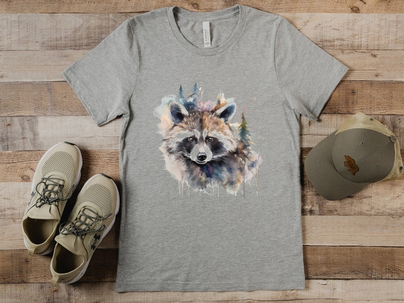 Puede incluir: Una camiseta gris con un dise&ntilde;o de mapache en acuarela y elementos forestales. La camiseta se combina con zapatillas de deporte beige y una gorra gris con un parche marr&oacute;n. Los art&iacute;culos est&aacute;n colocados sobre una superficie de madera.