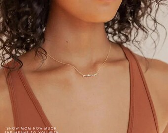 May include: A gold necklace with the word "mama" written in cursive script. The necklace is worn by a person with brown skin and dark curly hair.