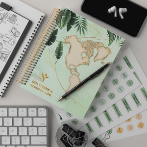 May include: A green and brown spiral bound journal with a world map design and the text "Royal Serenity, Cultivate a Blissful Life". The journal is on a white surface with a black pen, a silver keyboard, and a clear glass jar with black paper clips.