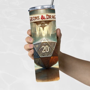 Puede incluir: Un vaso de acero inoxidable con pajita, con un dise&ntilde;o de fantas&iacute;a. El vaso muestra el texto "Dungeons & Dragons" y una ilustraci&oacute;n detallada de un dado de veinte caras con el n&uacute;mero 20. El dise&ntilde;o incluye un paisaje urbano y un mapa.