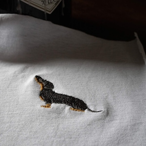 May include: A white t-shirt with an embroidered black dachshund. The dachshund is facing right and has a long body and short legs.
