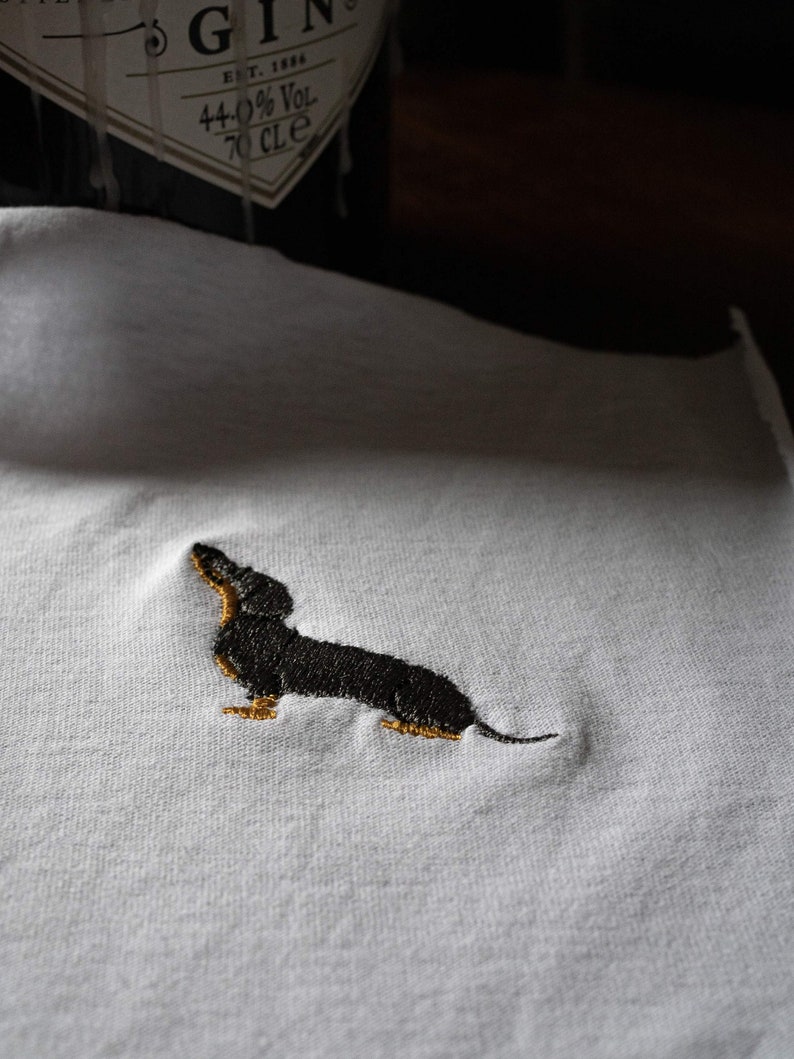 May include: A white t-shirt with an embroidered black dachshund. The dachshund is facing right and has a long body and short legs.