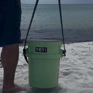 May include: A light green YETI bucket with a black handle is held by a person on a sandy beach. The bucket has a rectangular black label with the brand name. The background shows the ocean and a cloudy sky.