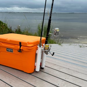 May include: Orange Yeti cooler with a white rod holder attached to the side. Two fishing rods are secured in the holder. The cooler is sitting on a wooden deck.