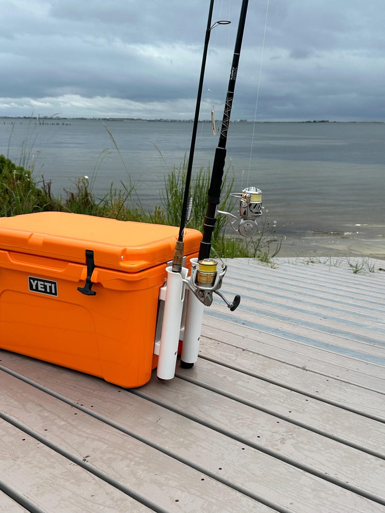 May include: Orange Yeti cooler with a white rod holder attached to the side. Two fishing rods are secured in the holder. The cooler is sitting on a wooden deck.