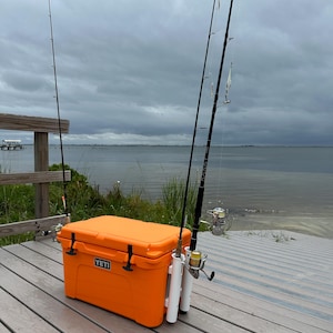 May include: Orange Yeti cooler with a white rod holder attached to the side. Three fishing rods are attached to the holder. The cooler is sitting on a wooden deck.
