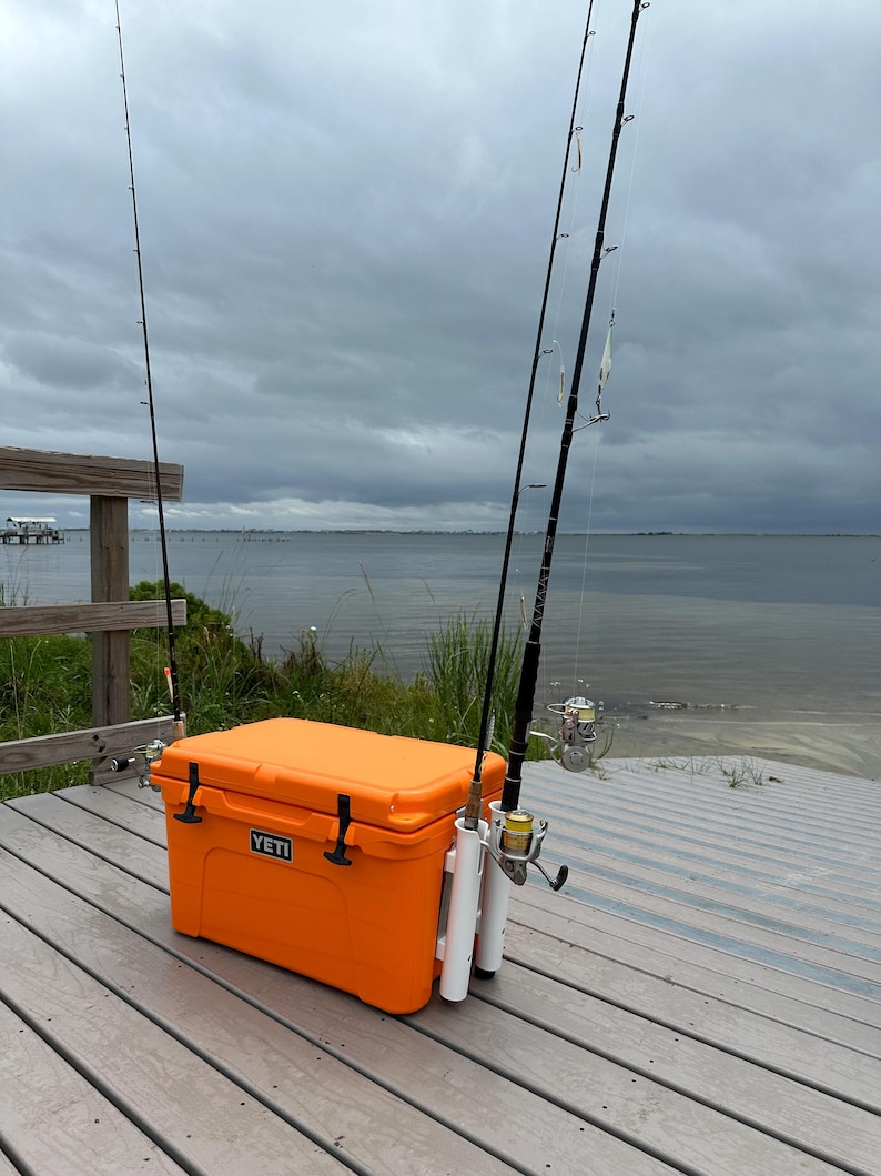 May include: Orange Yeti cooler with a white rod holder attached to the side. Three fishing rods are attached to the holder. The cooler is sitting on a wooden deck.