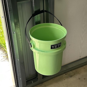 May include: A lime green Yeti bucket with a black handle hanging on a metal hook. The bucket is empty and has the Yeti logo on the side.
