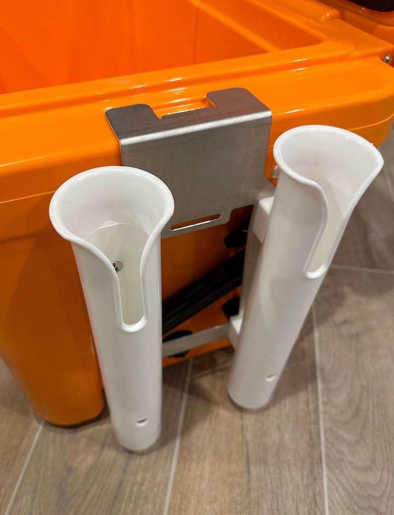 May include: Two white plastic rod holders mounted on a stainless steel bracket attached to an orange cooler.