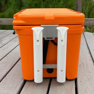 May include: Orange cooler with two white rod holders mounted on the side. The cooler is sitting on a wooden deck.