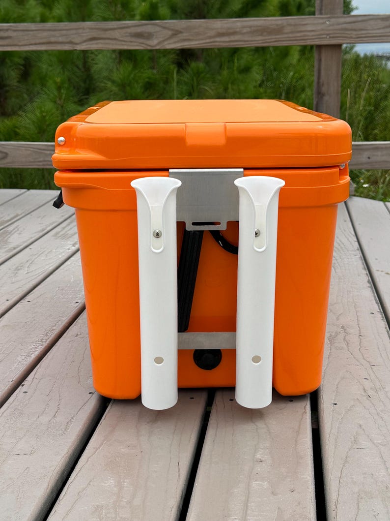 May include: Orange cooler with two white rod holders mounted on the side. The cooler is sitting on a wooden deck.