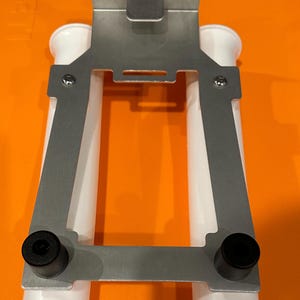 May include: A silver metal bracket with two white plastic tubes attached. The bracket has two black rubber feet on the bottom.