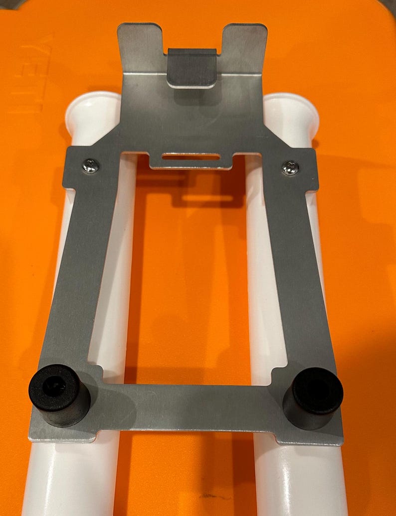 May include: A silver metal bracket with two white plastic tubes attached. The bracket has two black rubber feet on the bottom.