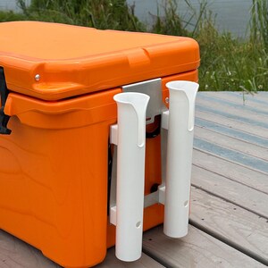 May include: Orange cooler with two white rod holders mounted on the side. The cooler is sitting on a wooden deck.