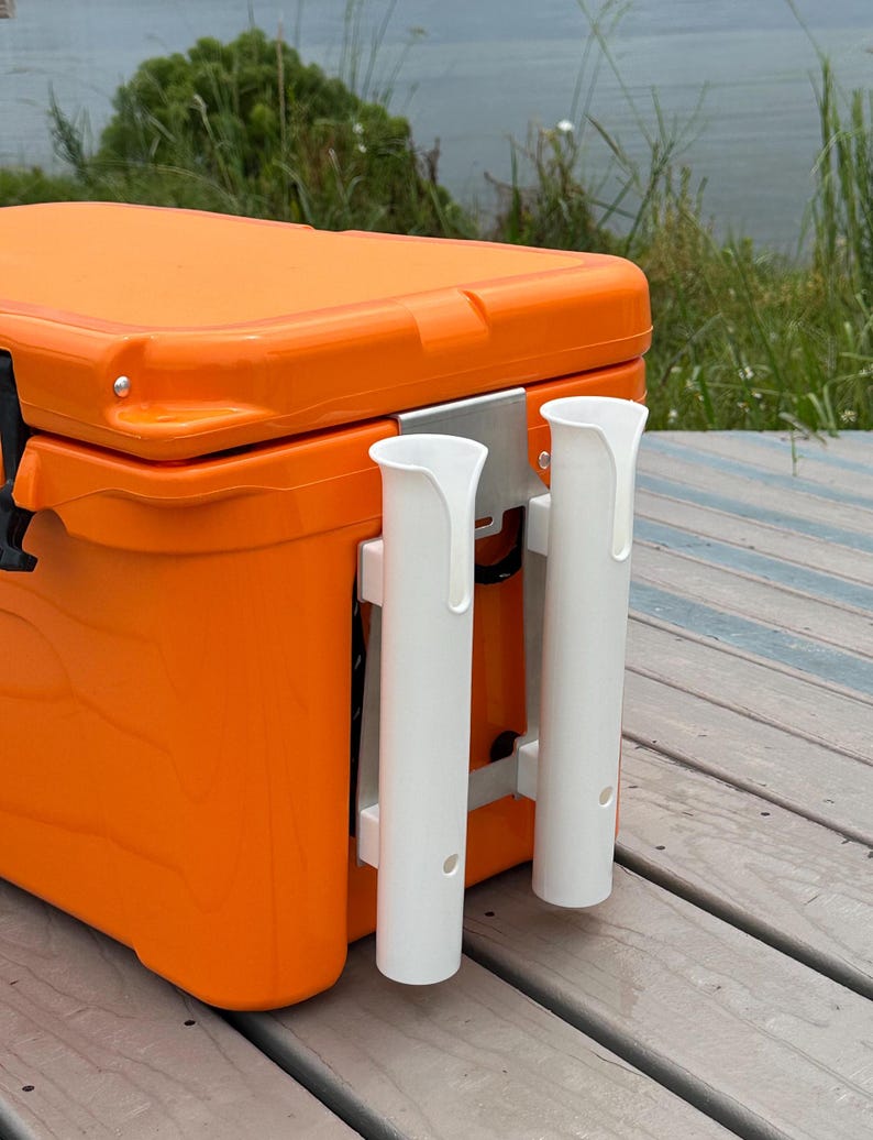 May include: Orange cooler with two white rod holders mounted on the side. The cooler is sitting on a wooden deck.