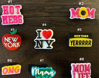 May include: Eight colorful shoe charms with various phrases and designs. The charms include "Hot Mess", "Mom", "New York", "I Love NY", "Yerrrrr", "OMG", "Mama", and "Mom Life".