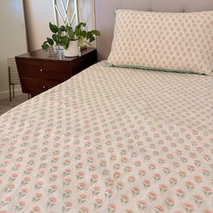 May include: A white bed sheet and matching pillowcase featuring a repeating pattern of small pink flowers with green stems and leaves. The bedding set has a light green trim along the edge of the pillowcase and is spread across a bed frame.
