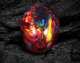 May include: A fiery red, black, and green opal gemstone with a smooth, oval shape. The opal is set against a dark, textured background.
