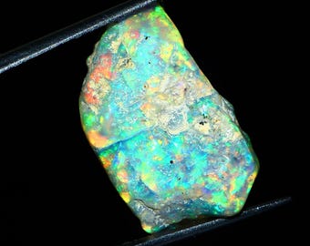 May include: A rough, iridescent opal gemstone with a green, blue, and orange play of color. The opal is held in tweezers against a black background.