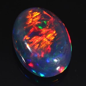 Pu&ograve; includere: Una pietra preziosa di opale nero con un bagliore iridescente rosso, arancione e verde. La pietra di forma ovale &egrave; incastonata in una montatura trasparente e lucida.