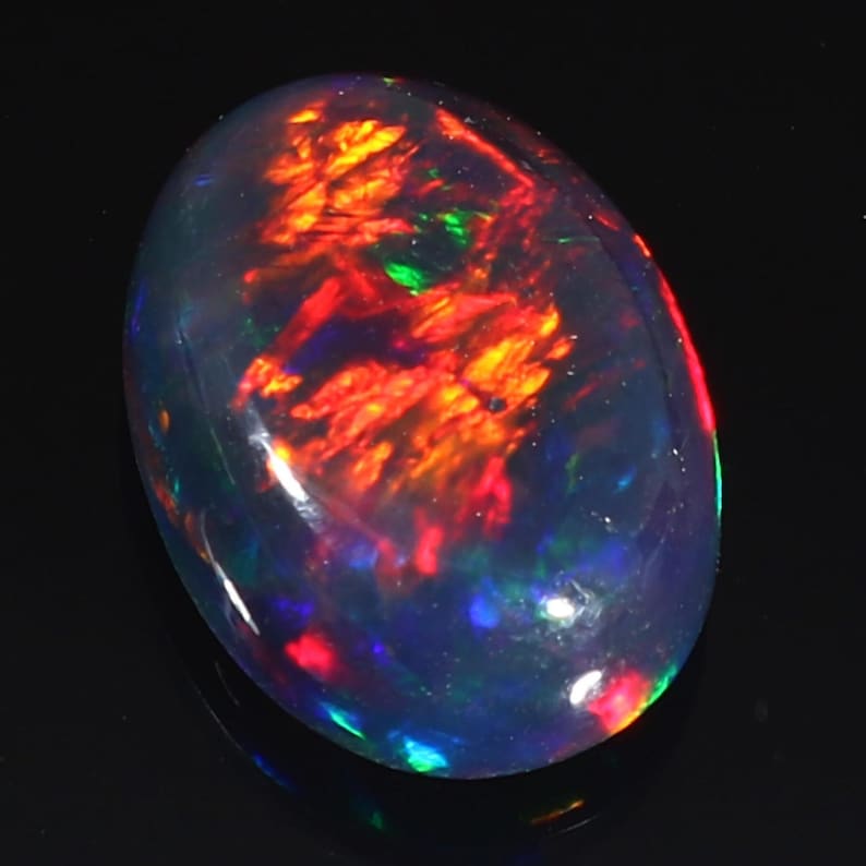 Pu&ograve; includere: Una pietra preziosa di opale nero con un bagliore iridescente rosso, arancione e verde. La pietra di forma ovale &egrave; incastonata in una montatura trasparente e lucida.