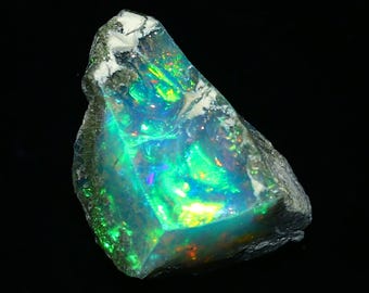 May include: A raw opal gemstone with a dark gray exterior and vibrant internal colors. The opal displays flashes of green, blue, and yellow, creating a shimmering effect. The stone is irregularly shaped, showcasing its natural form.