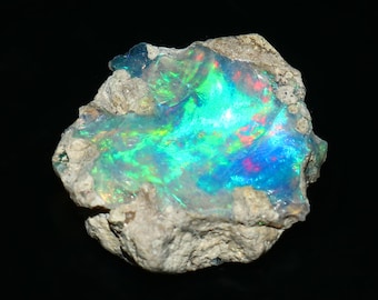 May include: A rough, uncut opal gemstone with a vibrant display of color. The opal's surface showcases a play of light with iridescent flashes of blue, green, and red. The stone is set in a matrix of gray rock.