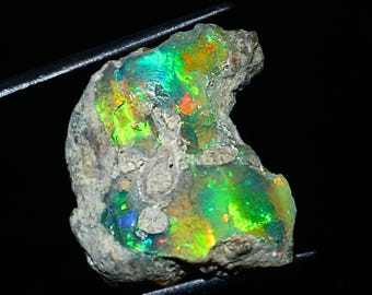 May include: A rough-cut opal gemstone with vibrant colors of green, orange, and blue. The opal is held by a pair of tweezers against a black background. The stone has a rough, textured exterior.